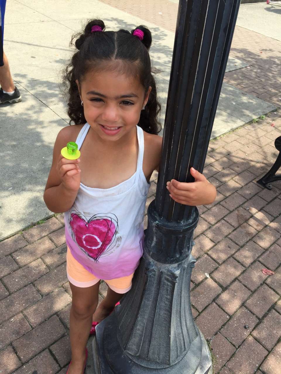 girl with 2 ponytails holding streetlamp with green ring pop and desigual kids heart tank top