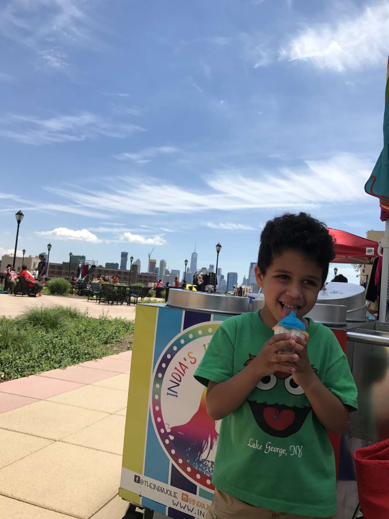 Litle boy with oscar the ground tshirt standing in front of India's Italian Ice cart smiling holidng a blue Italian Ice in his hand with views of NYC in the background