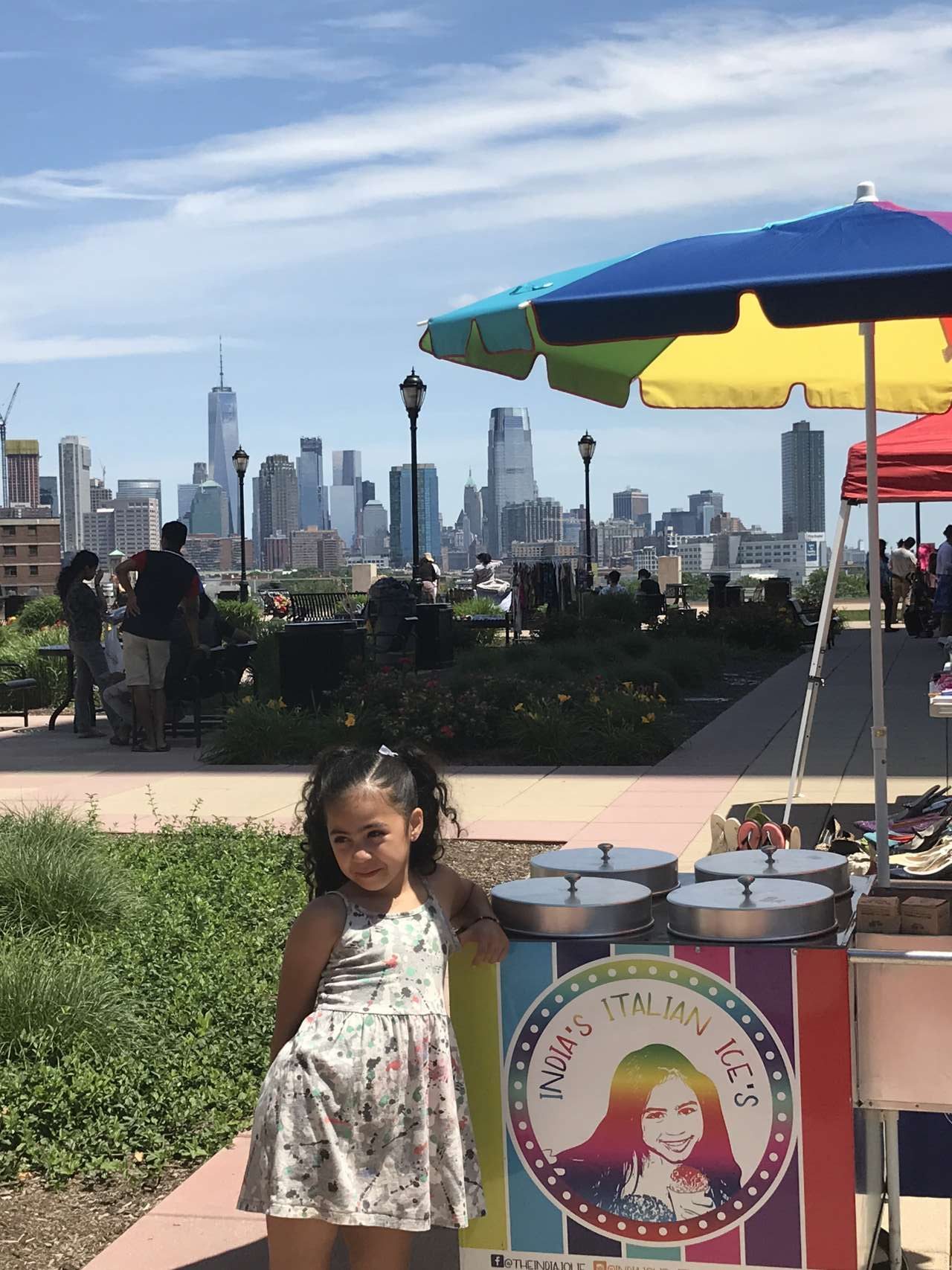 girl leaning on India's Italian Ice Cart with view of NYC in the background on a promenade