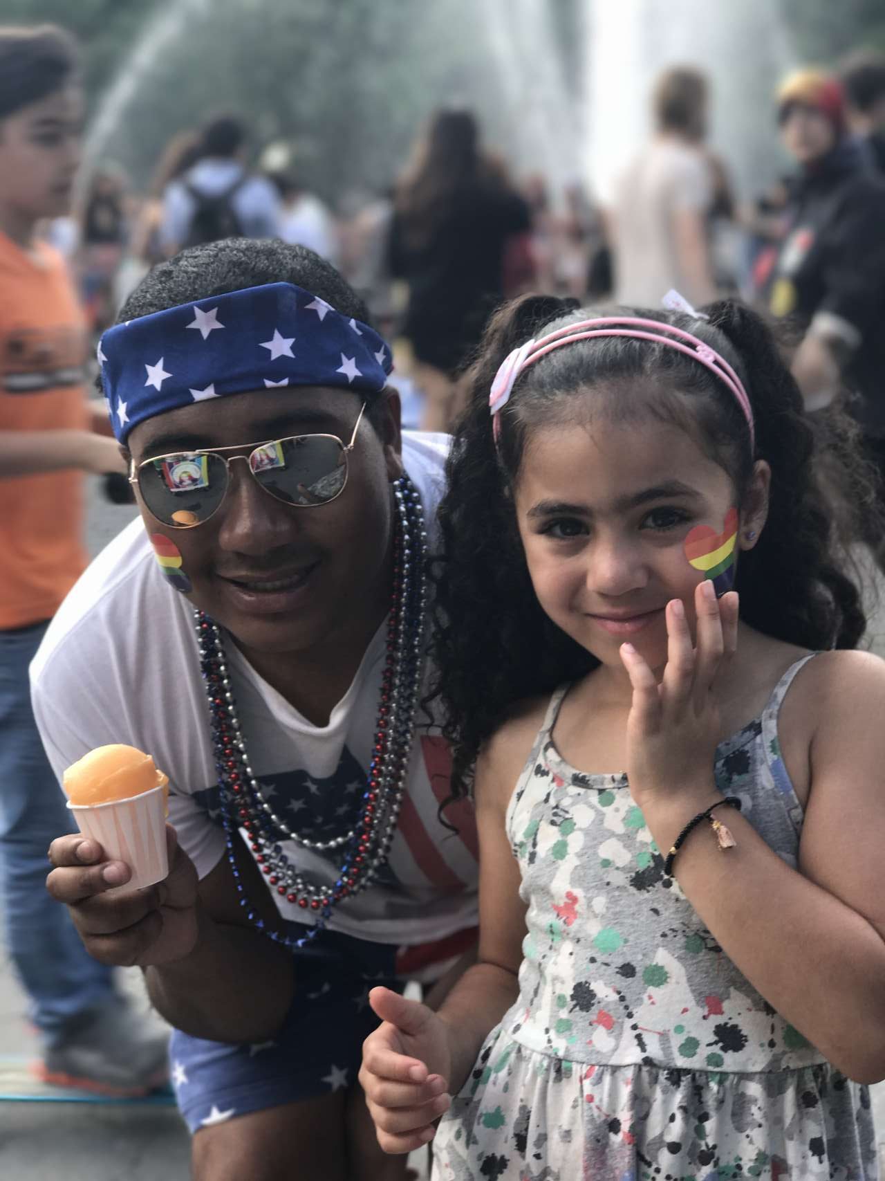 Man with bandana, sunglasses and rainbow heart sticker on face holding a mango italian ice next to girl, India Jolie with rainbow heart sticker on face wearing printed dress with fountain in background