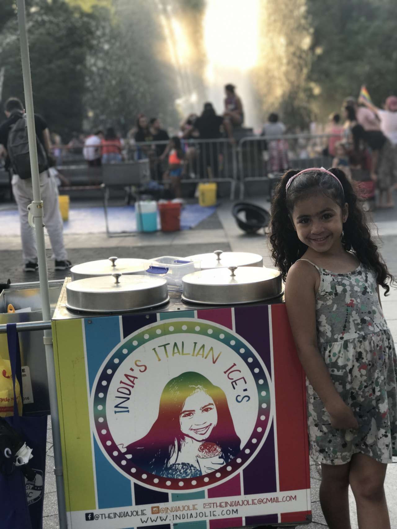 girl India Jolie in printed dress standing next to her rainbow Italian Ice cart in Washington Square Park