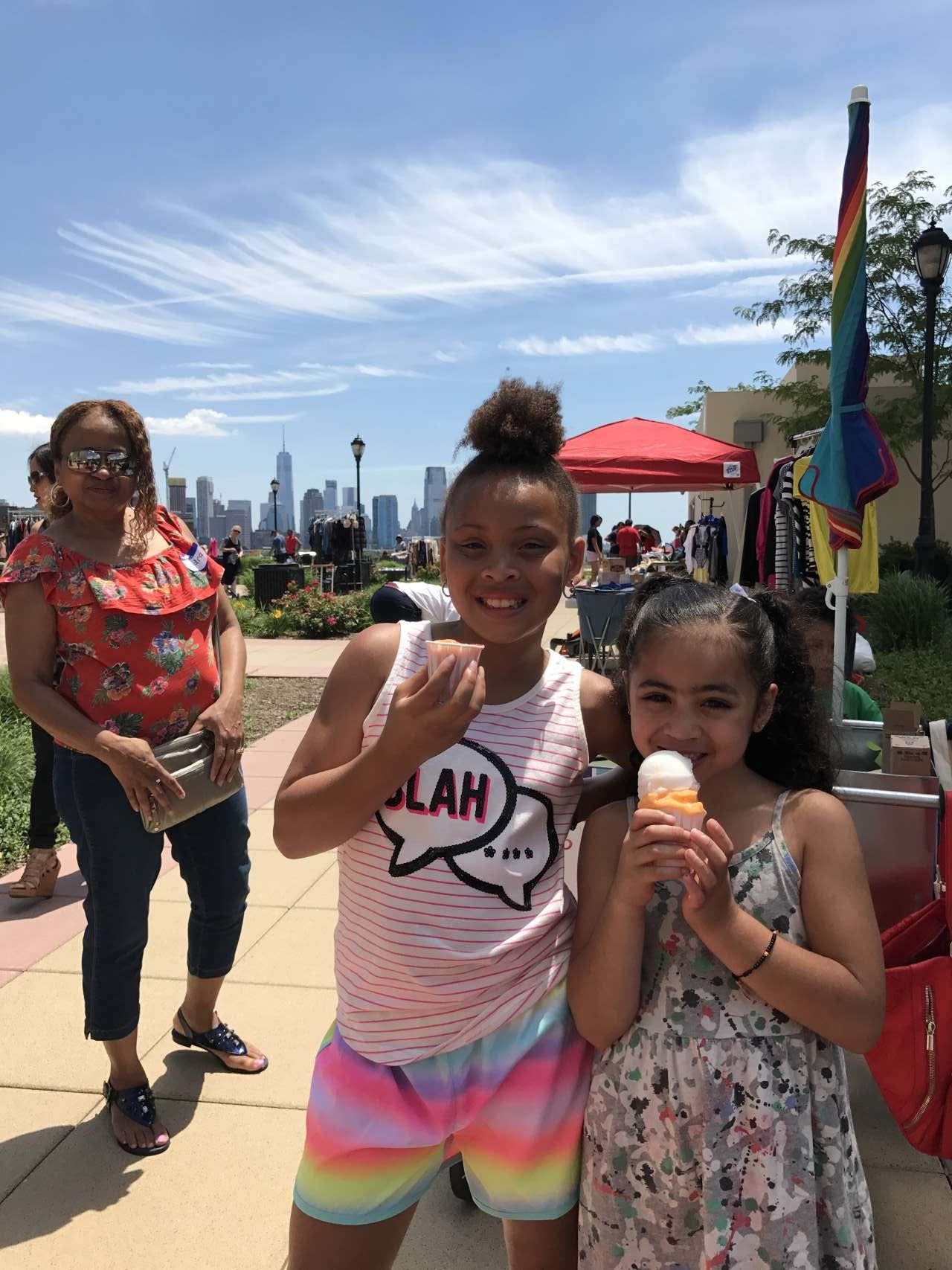 2 girls eating Italian Ice with brightly printed clothing on a summer day with NYC views in the background 