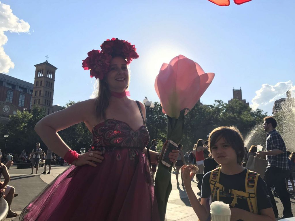 Women holding oversized dramaatic pink flower wearing flower headband and pink dress next to boy eating italian ice in park
