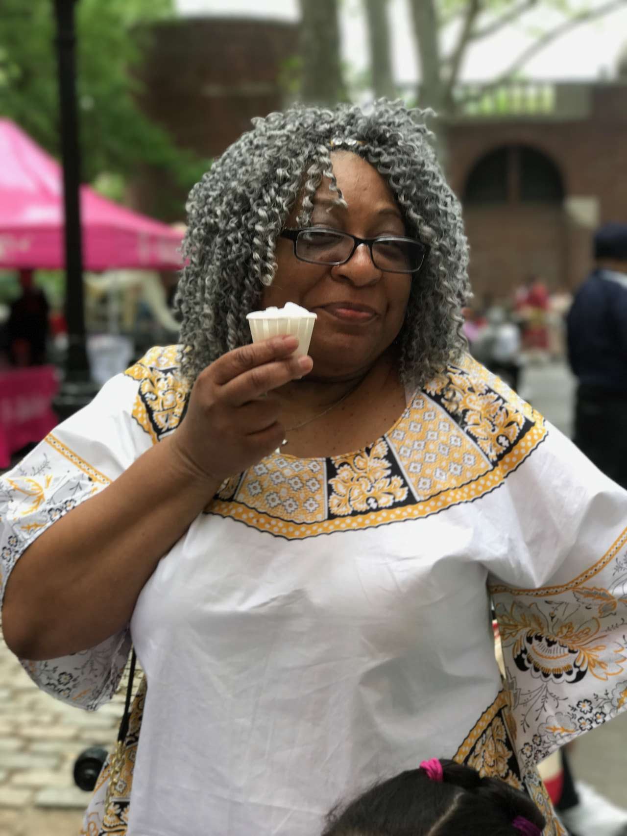 older woman smiling with grey spiral curls in white african dress with gold and black trim smiling and holding lemon italian ice