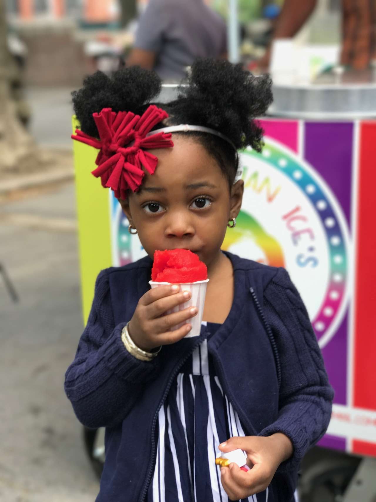 little girl with afro puff ponyails holding cherry red italian ice and wearing cherry red headband in front of rainbow italian ice cart